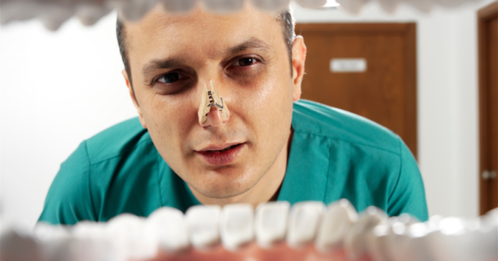 dentist looking into mouth while wearing a clothes pin on his nose because of the patients bad breath